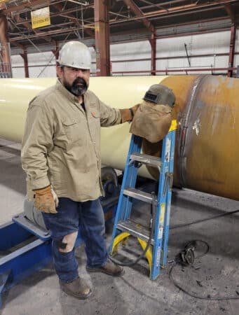 Mario Rocha (welder) standing next to steel pipe fitting in fabrication shop.