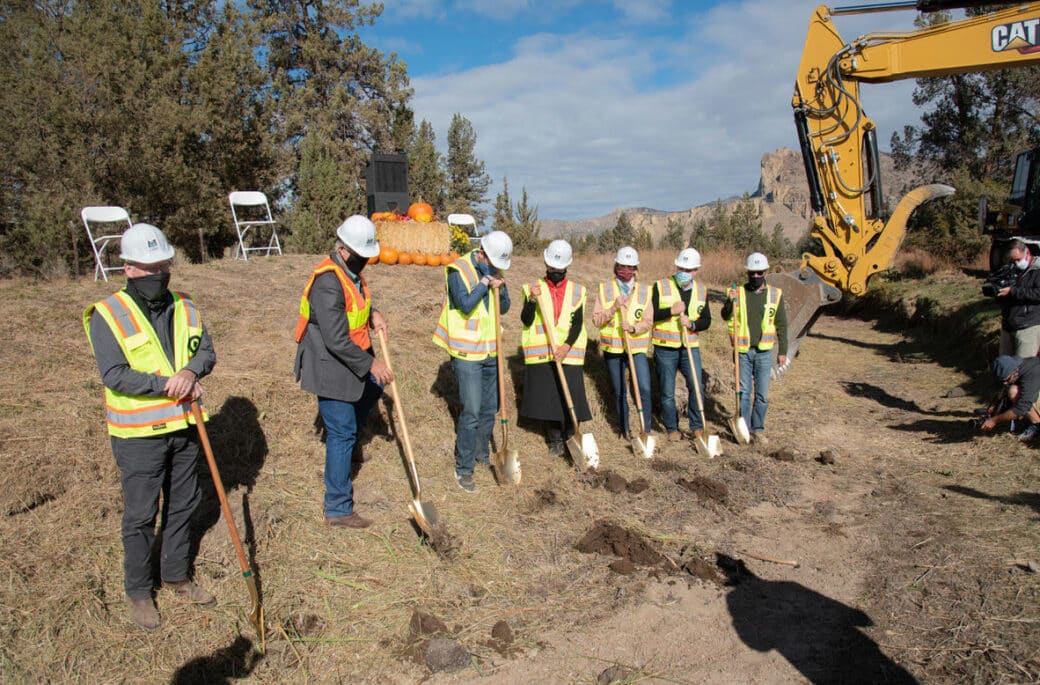 6 people in hard hats and vests with shovels standing in rural area with crane in background