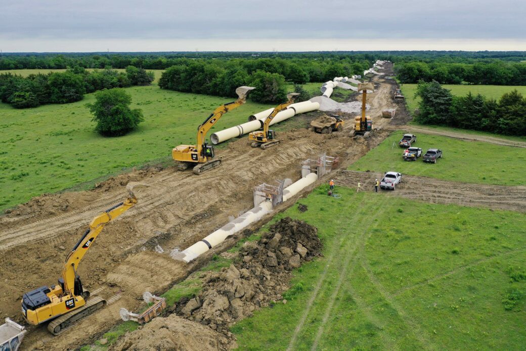 Large diameter steel pipe being installed by cranes into trench dug into grassy landscape