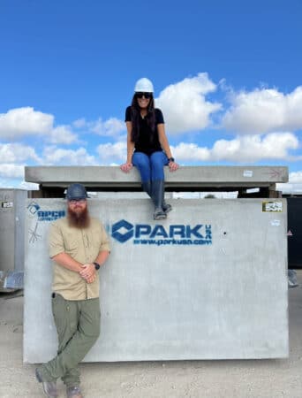 Woman in hard hat sitting on concrete vault, man in hardhat stands on the ground leaning against vault